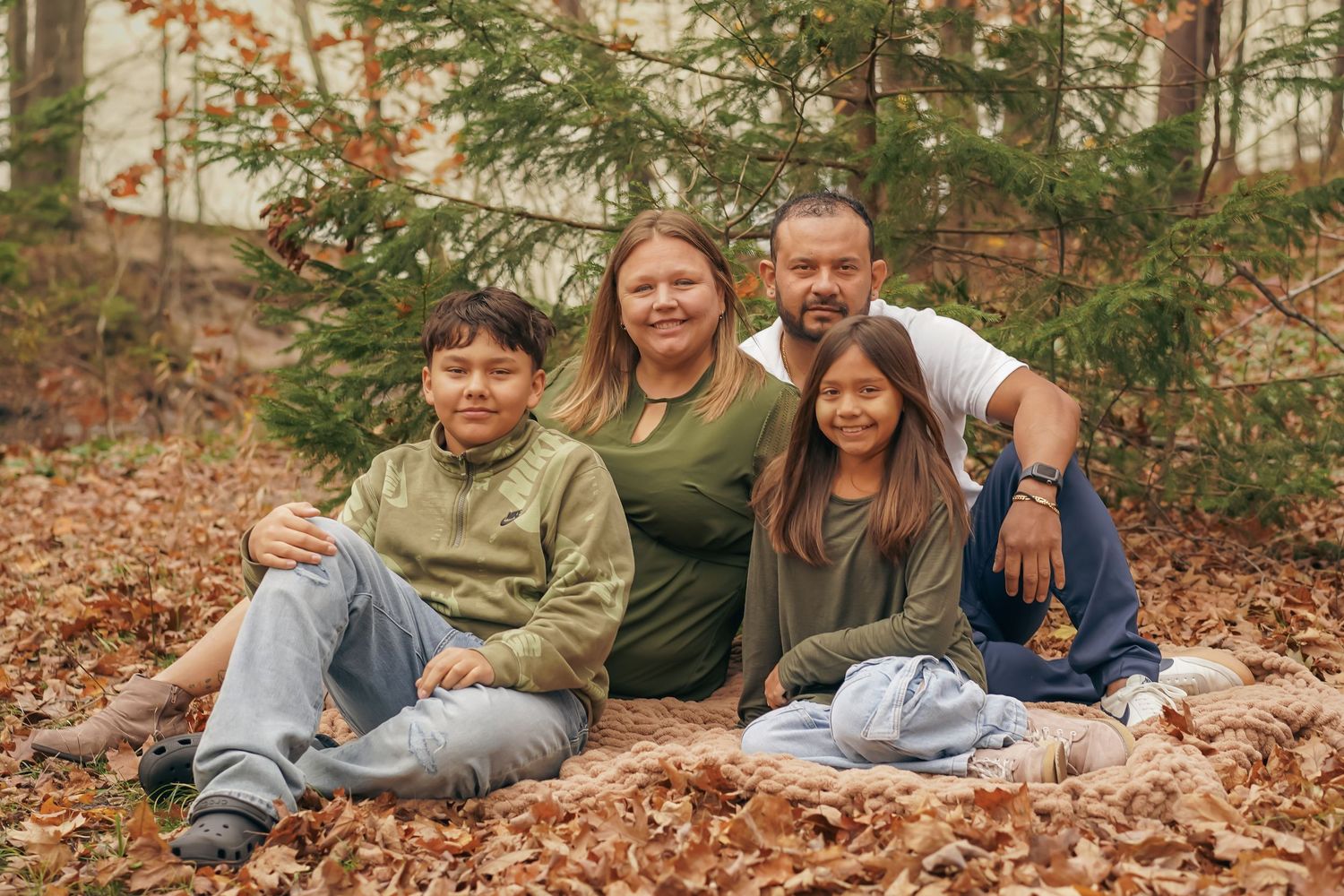 Family of four sits together on fallen autumn leaves in front of evergreen trees during outdoor photo session.