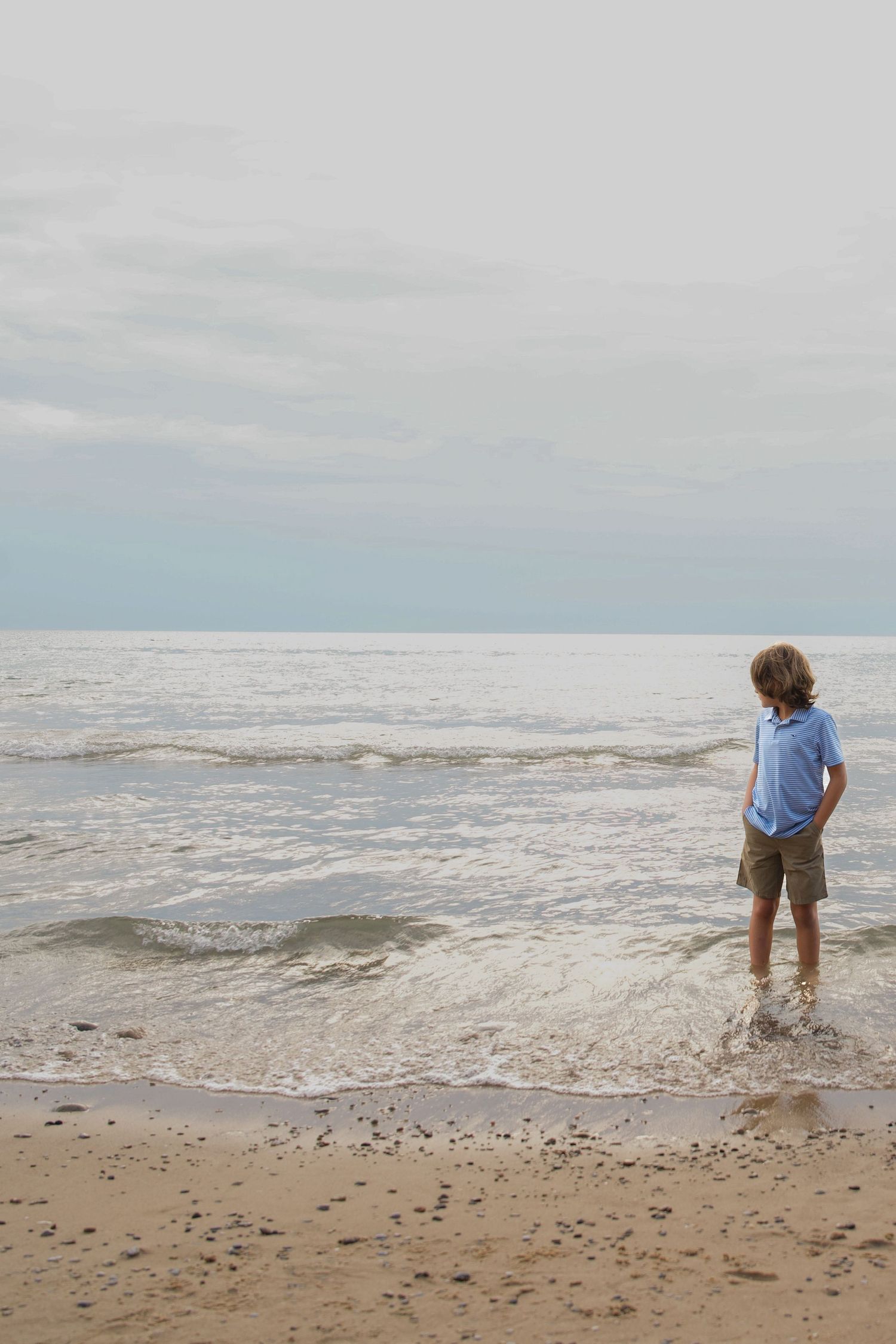 Person stands at the edge of the ocean waves on a sandy beach under an overcast sky.
