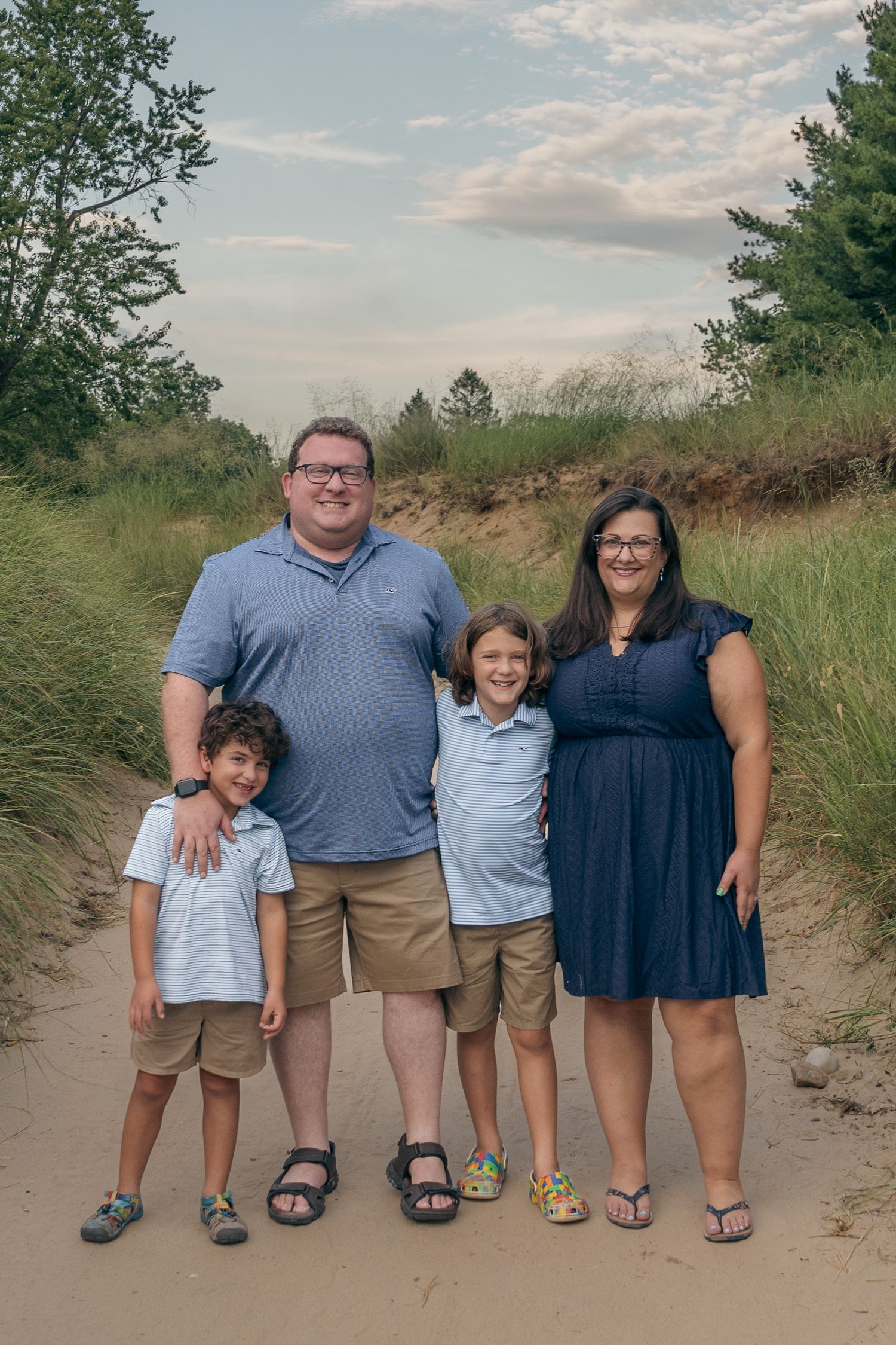 A family poses together on a sandy trail surrounded by green vegetation on a summer evening.