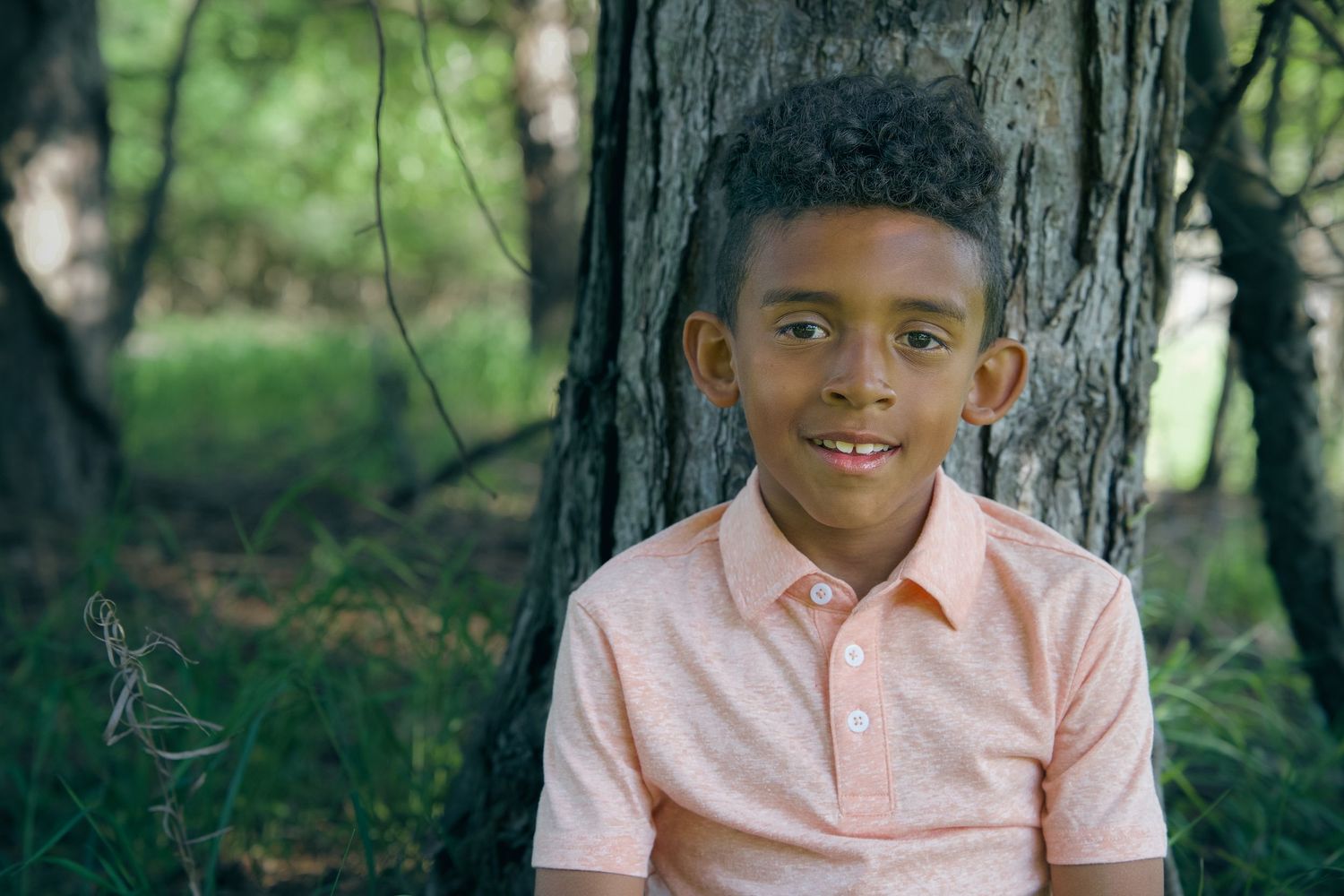 A young child in a peach-colored polo shirt smiles while sitting against a tree trunk in a wooded outdoor setting.