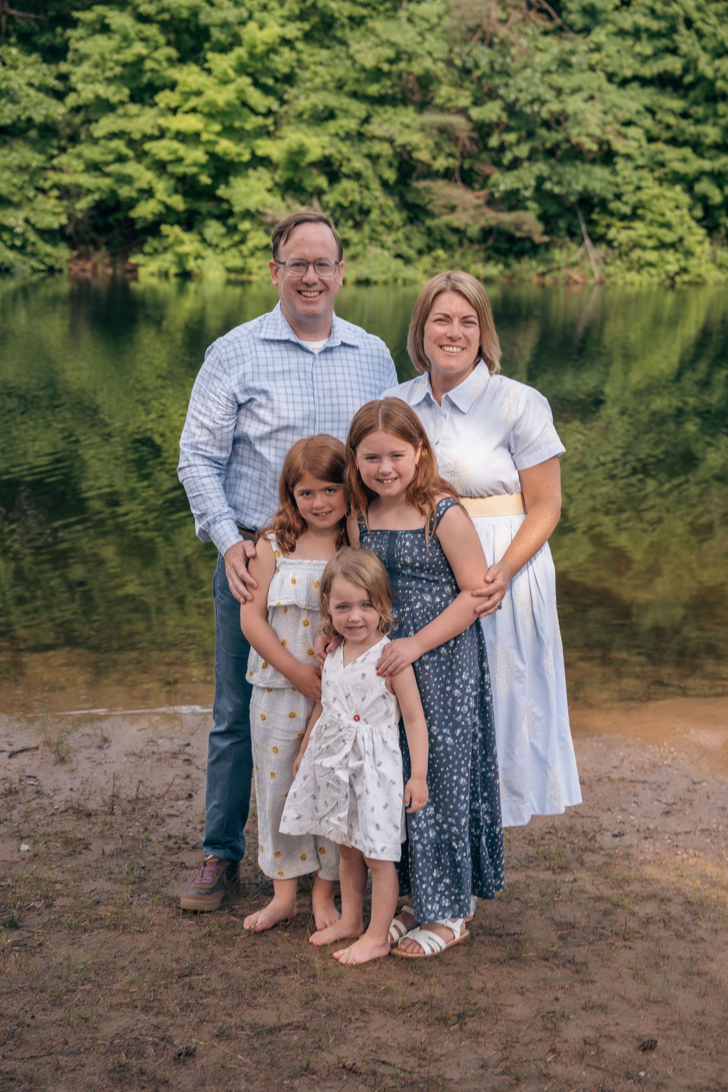 Happy family of five posing outdoors near a serene lake surrounded by lush green trees.
