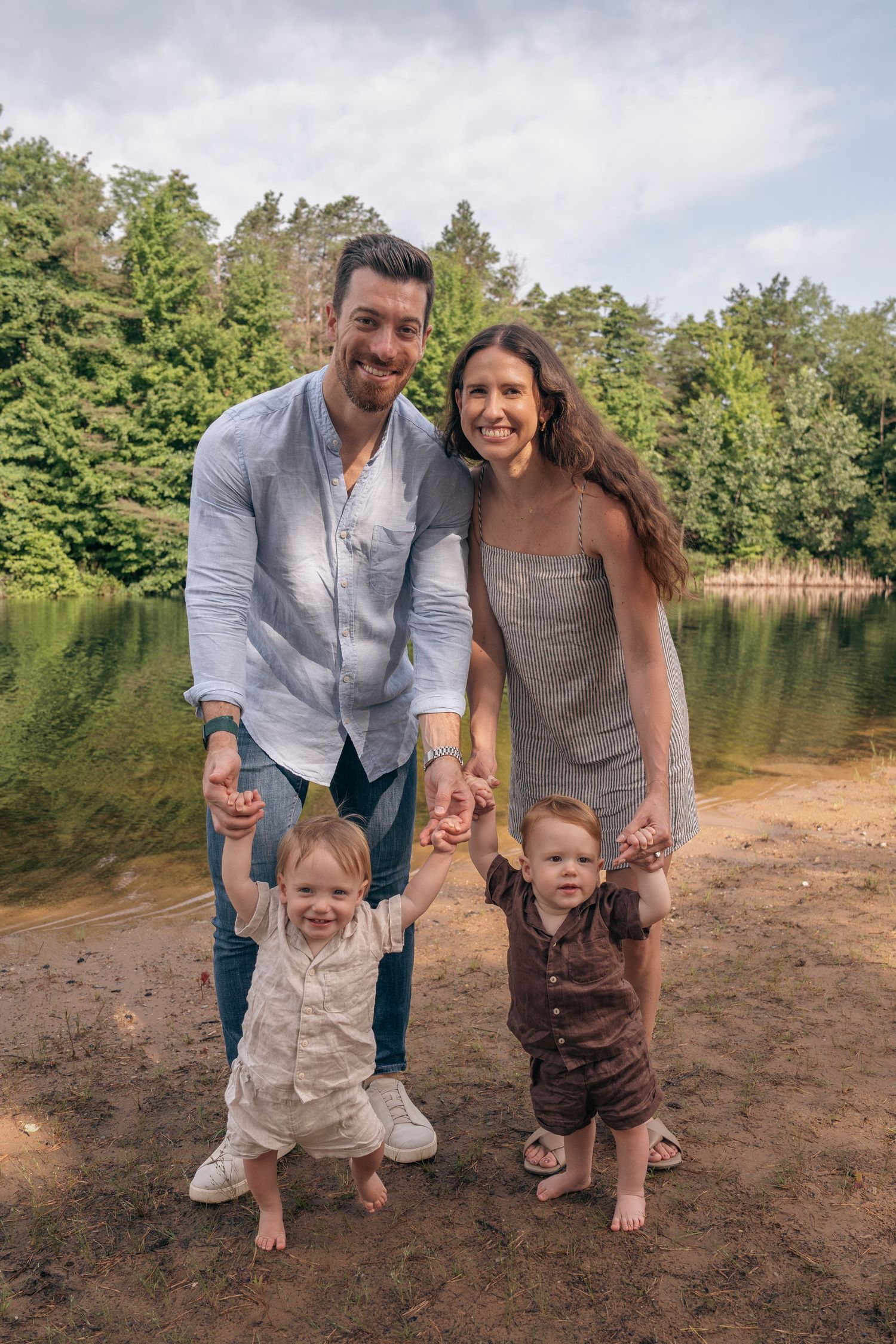 Happy family of four posing by a lake, parents holding toddlers hands, surrounded by lush green trees.