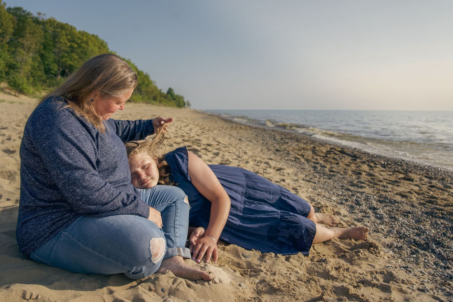 Person in denim sits thoughtfully on a rocky beach at sunset while looking out towards the ocean horizon.