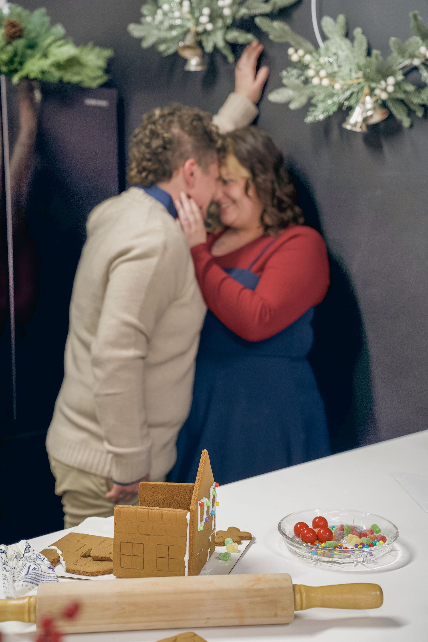 Two people share a romantic moment at a dining table with a gingerbread house and holiday decorations.