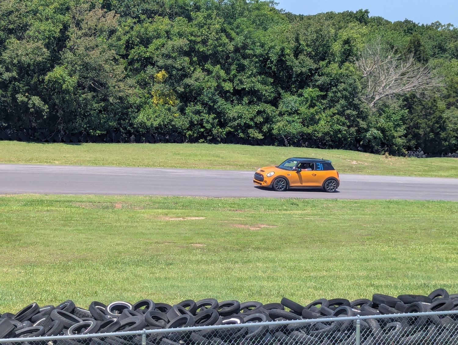 Bright orange sports car driving on a race track with green trees and tire barriers in the background.