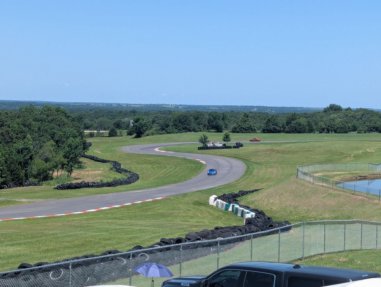 Aerial view of a winding racetrack with green landscaping, trees and a small pond on a sunny day.