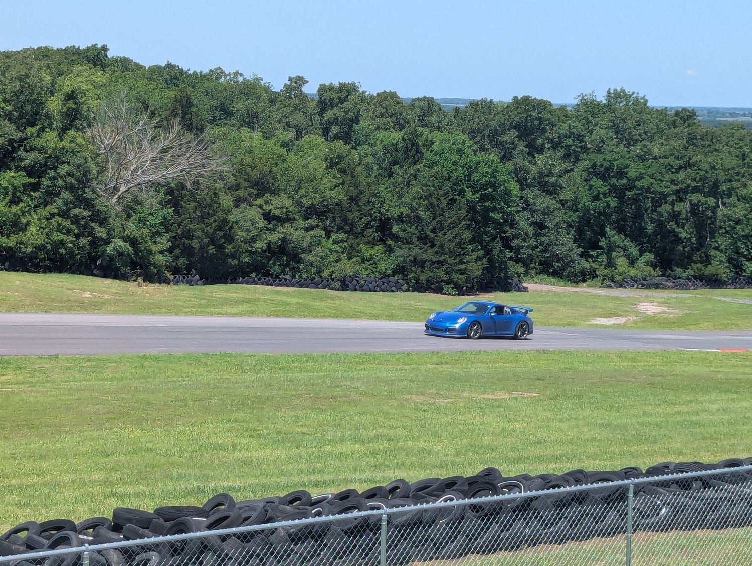 Blue sports car drives on a race track bordered by green grass and trees on a sunny day.