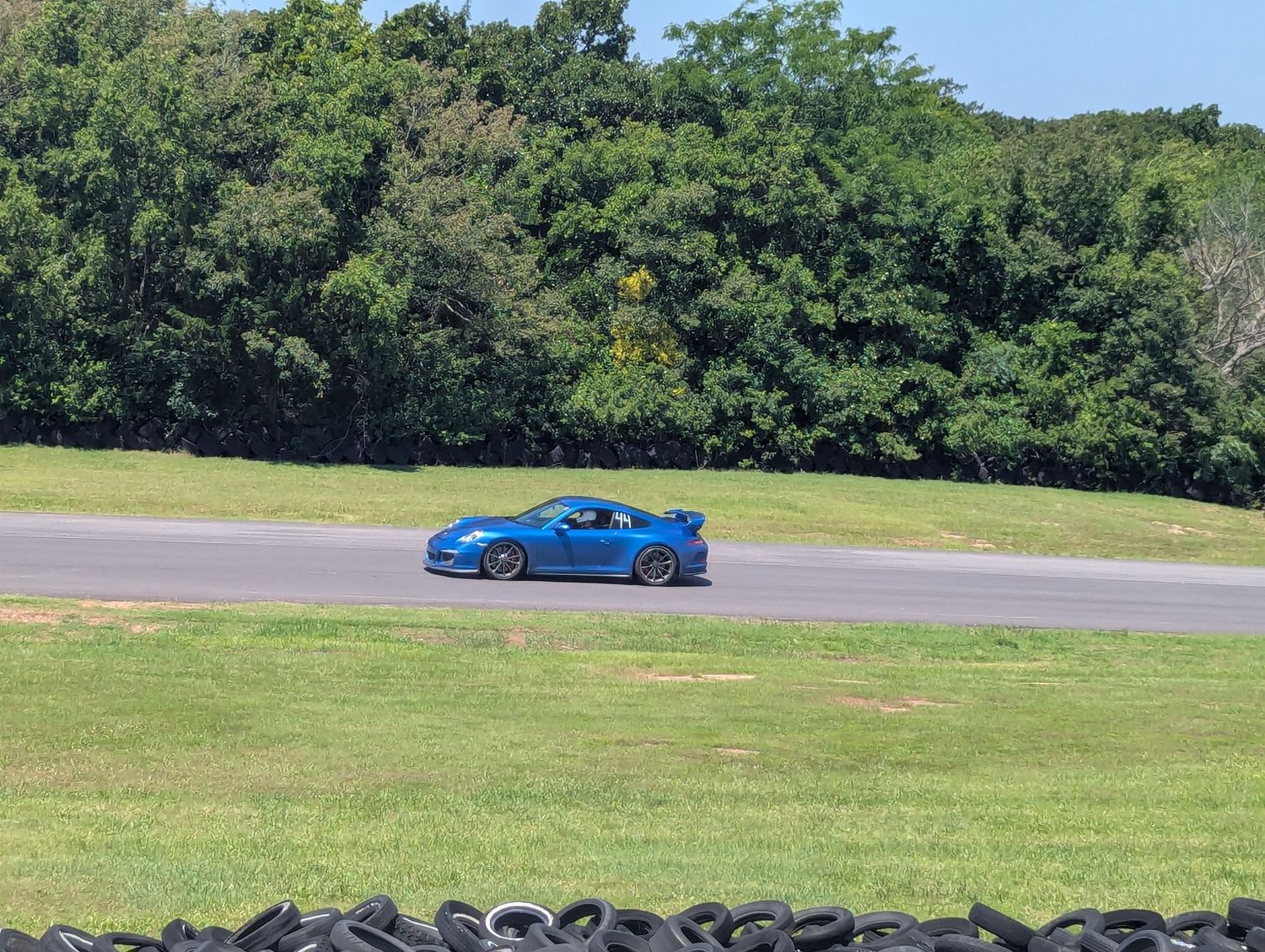 Blue sports car speeds along a race track bordered by grass and trees on a sunny day.