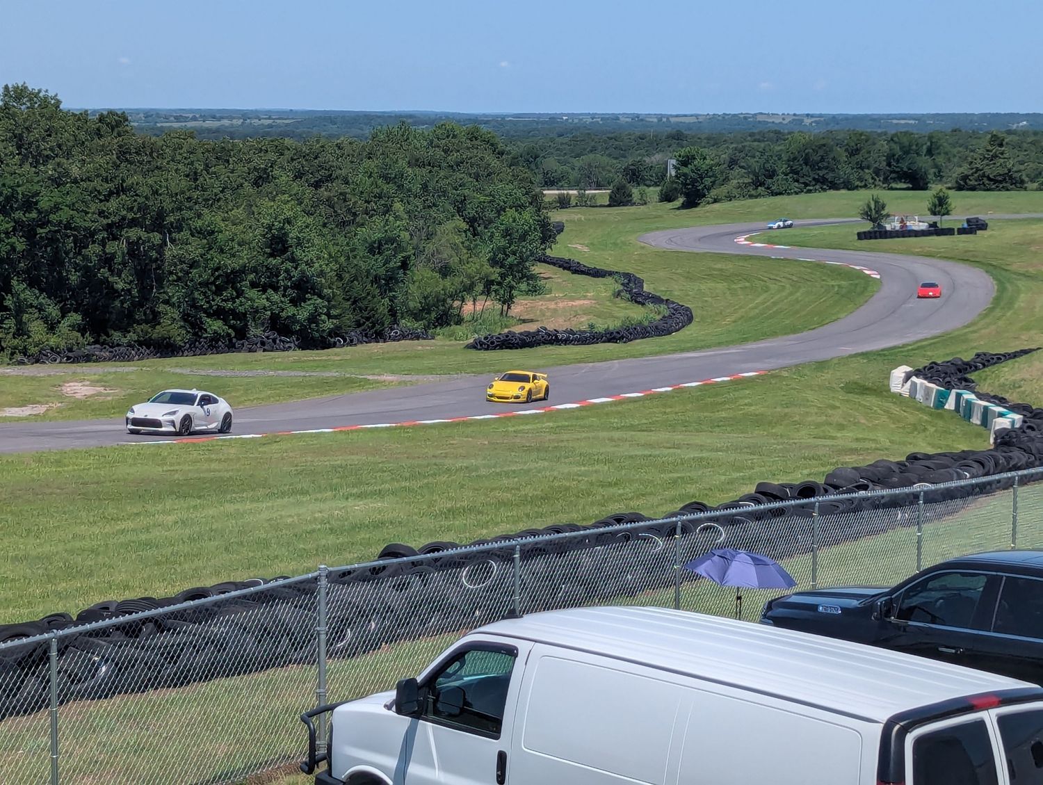Elevated view of a winding race track with multiple cars competing against a forested landscape.