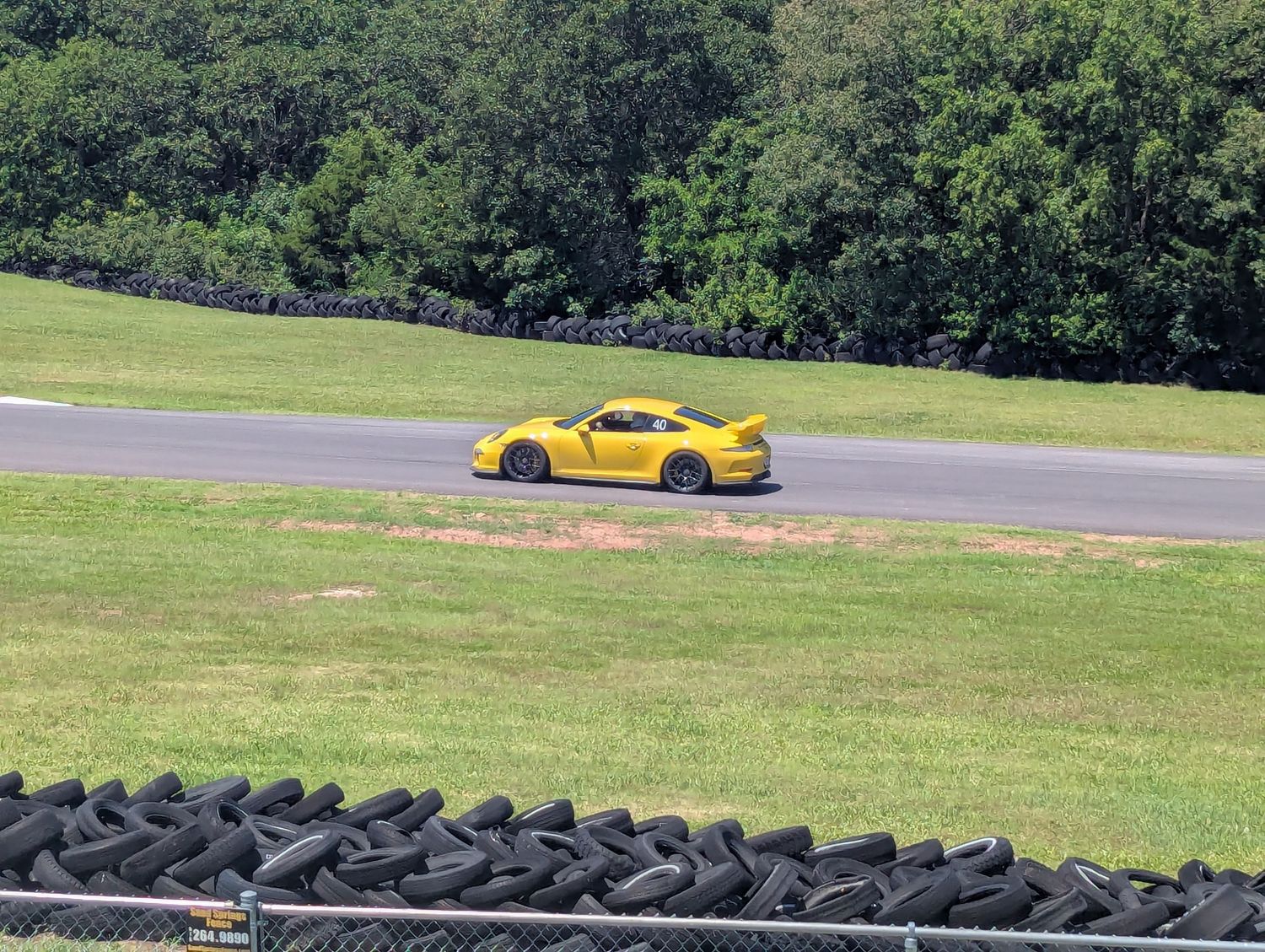 Row of used black tires laid out on grass near a racing track with trees in background.