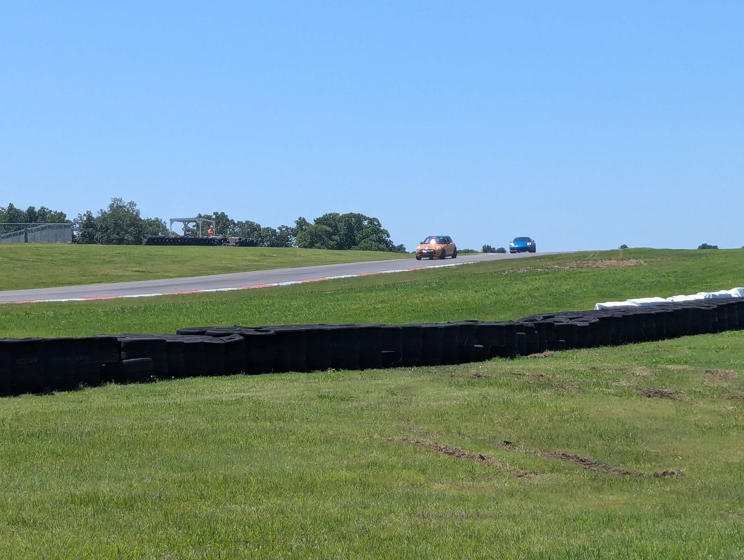 Cars racing on a sunny racetrack with green grass and trees in the background.