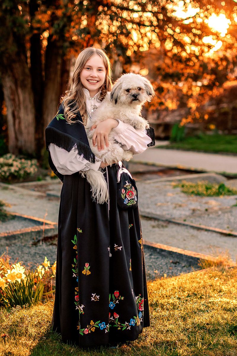 Konfirmant i Jelsabunad med hunden sin i armene i solnedgang på Eiganes Gravlund i Stavanger.