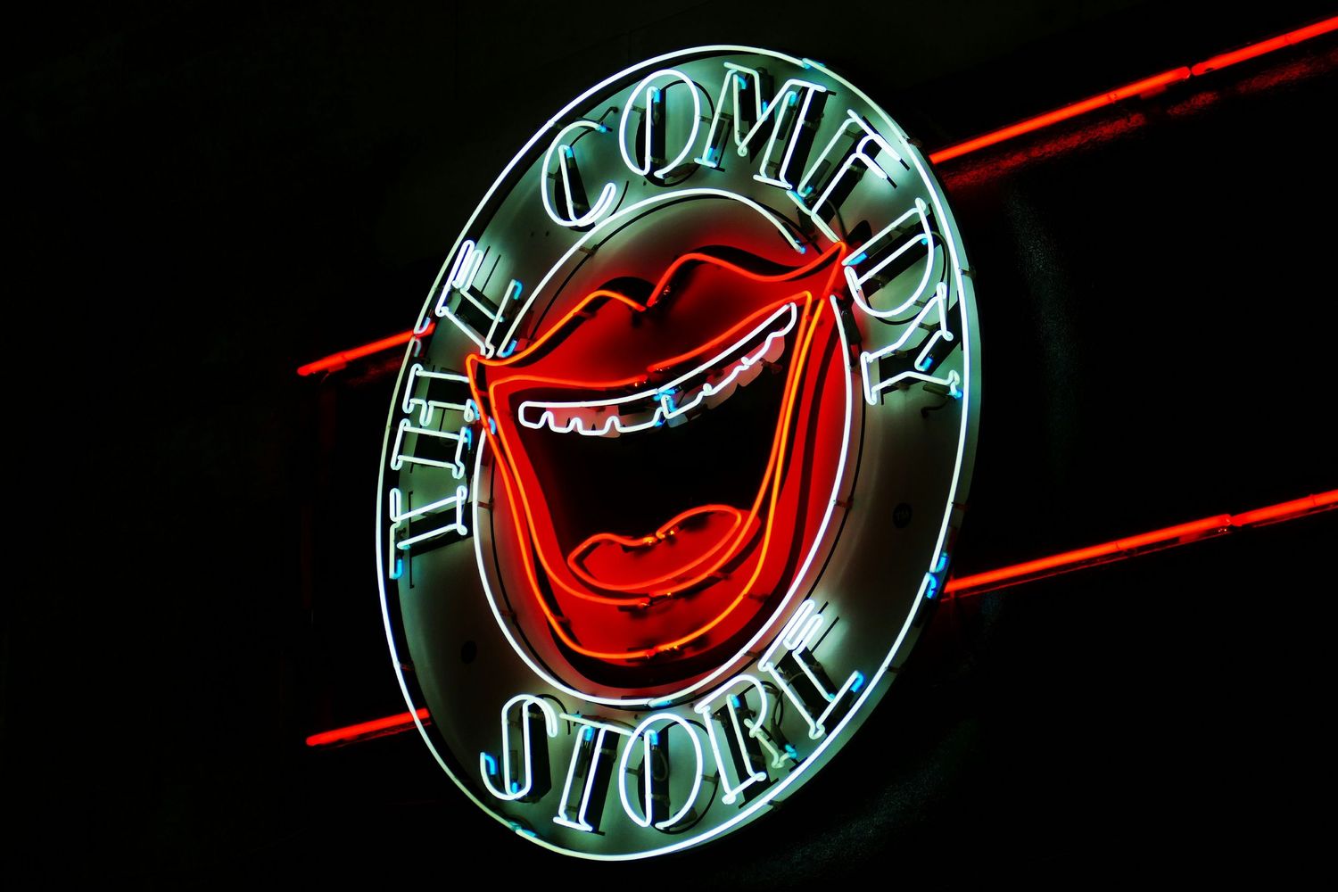 Neon sign featuring a glowing Comedy Store logo with a laughing mouth design and red accent lines.