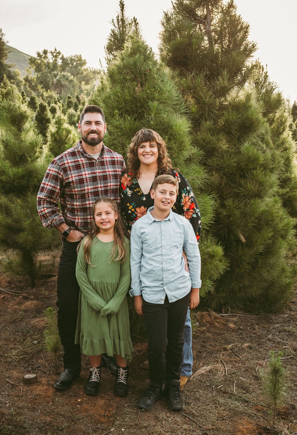 A family poses together in front of tall evergreen trees at a Christmas tree farm during golden hour.