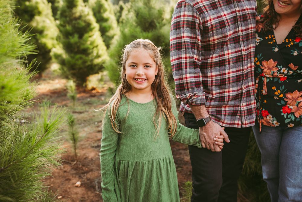 A child in a green dress stands among evergreen trees at a Christmas tree farm while holding someone's hand.