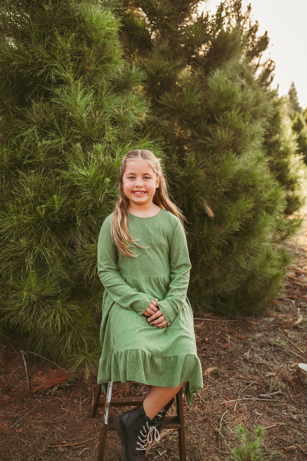 A child in a green dress and black boots stands next to an evergreen tree during golden hour.