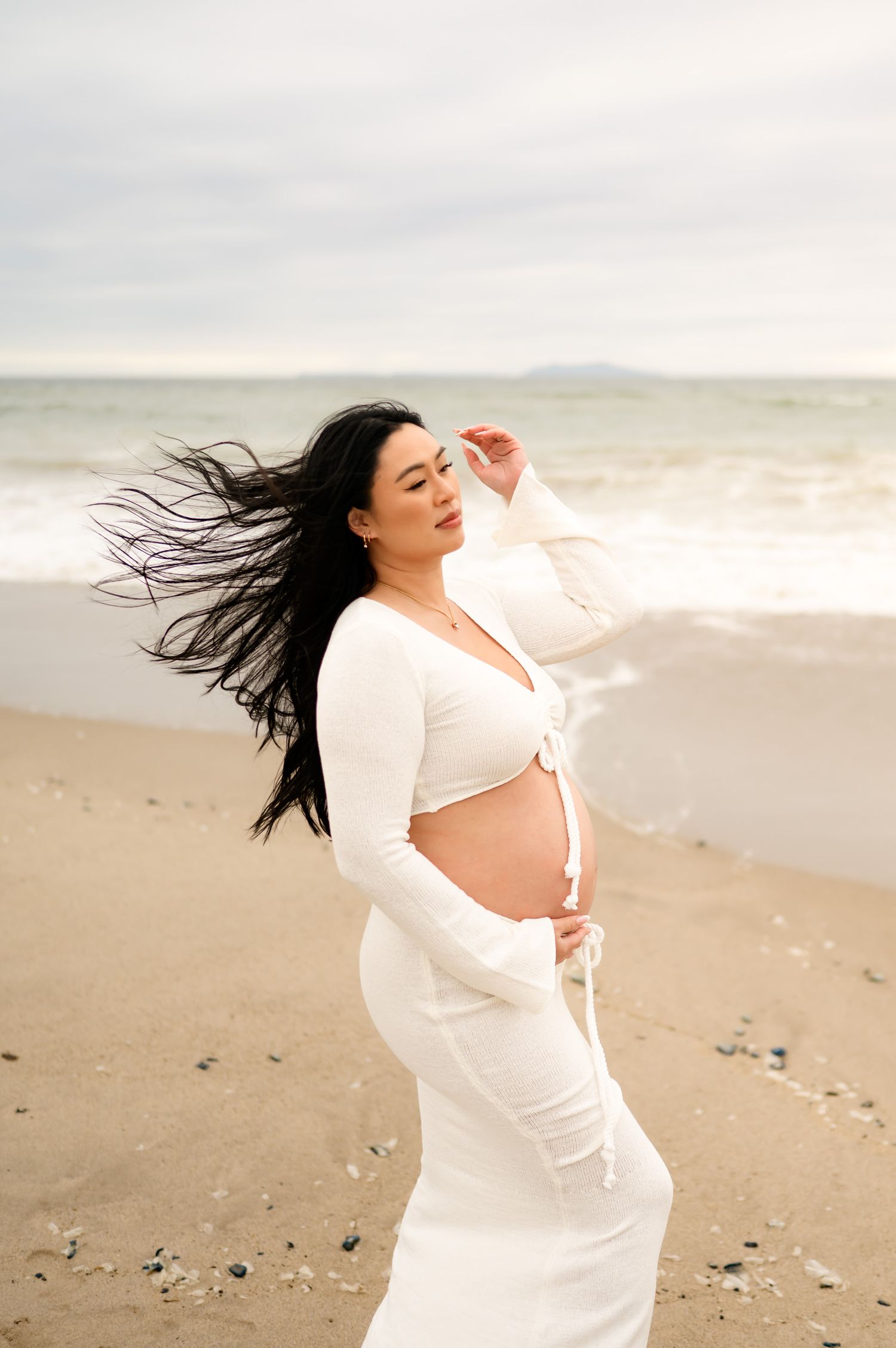 woman on in white two piece dress holds bare belly during her Ventura Beach maternity photoshoot while on her Santa Barbara babymoon