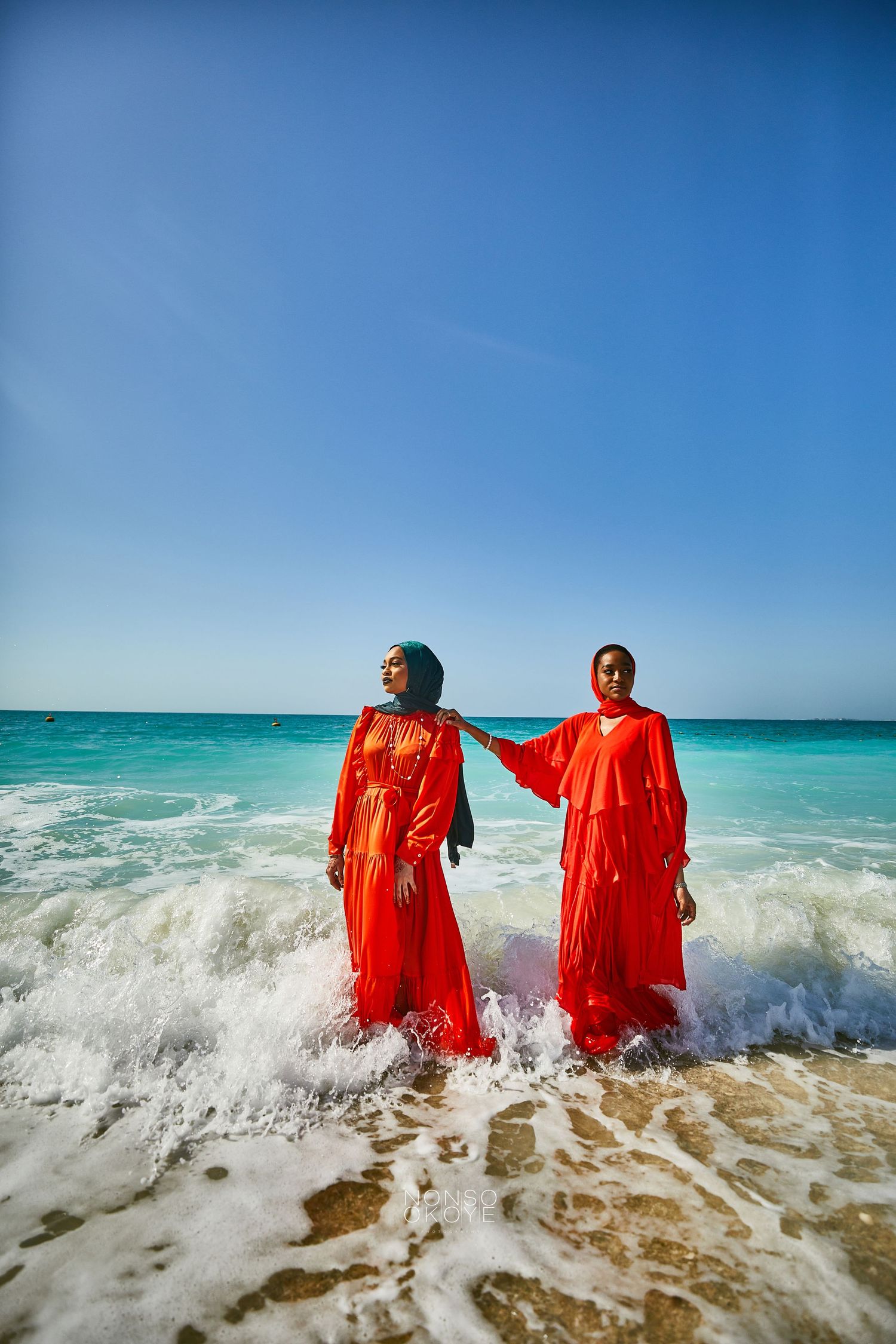 Two people in flowing red dresses standing in turquoise ocean waves at the beach.