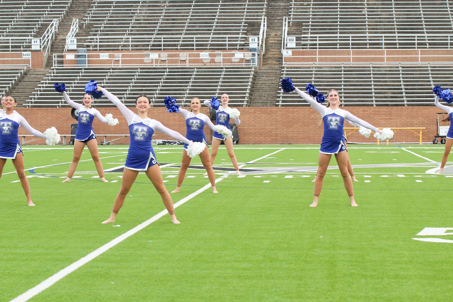 Cheerleaders in blue uniforms perform a synchronized dance routine on a football field with empty stadium bleachers.