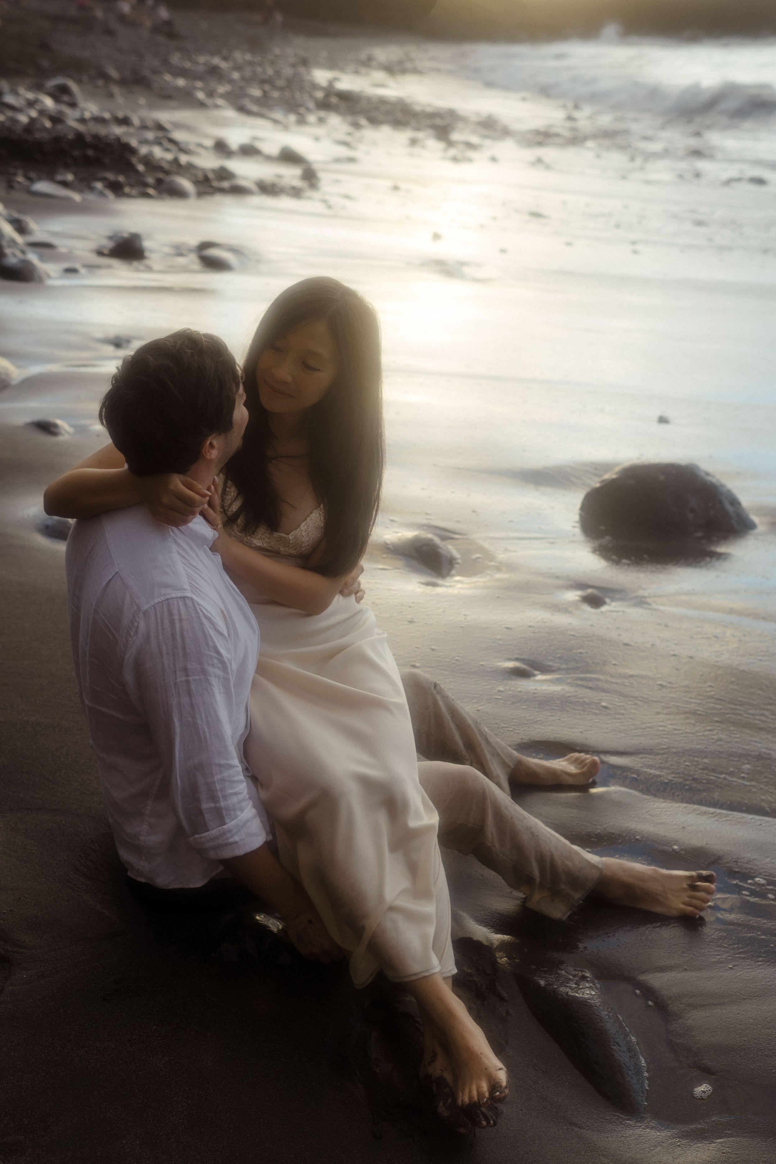 Romantic couple sitting together on a beach at sunset, woman in white dress embracing man.