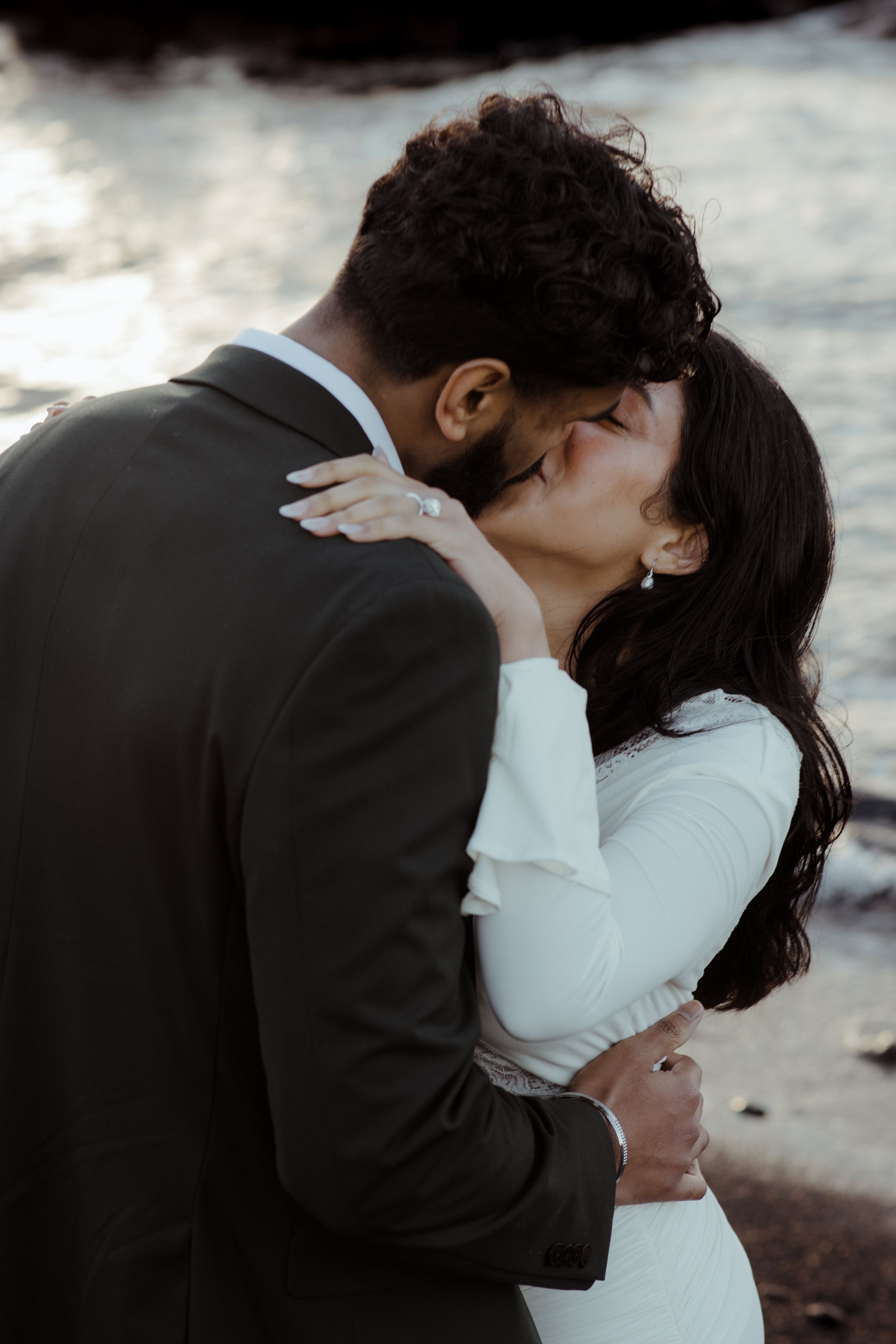 A couple shares a romantic kiss, the bride in a white dress and groom in a dark suit, by a shimmering waterside backdrop.