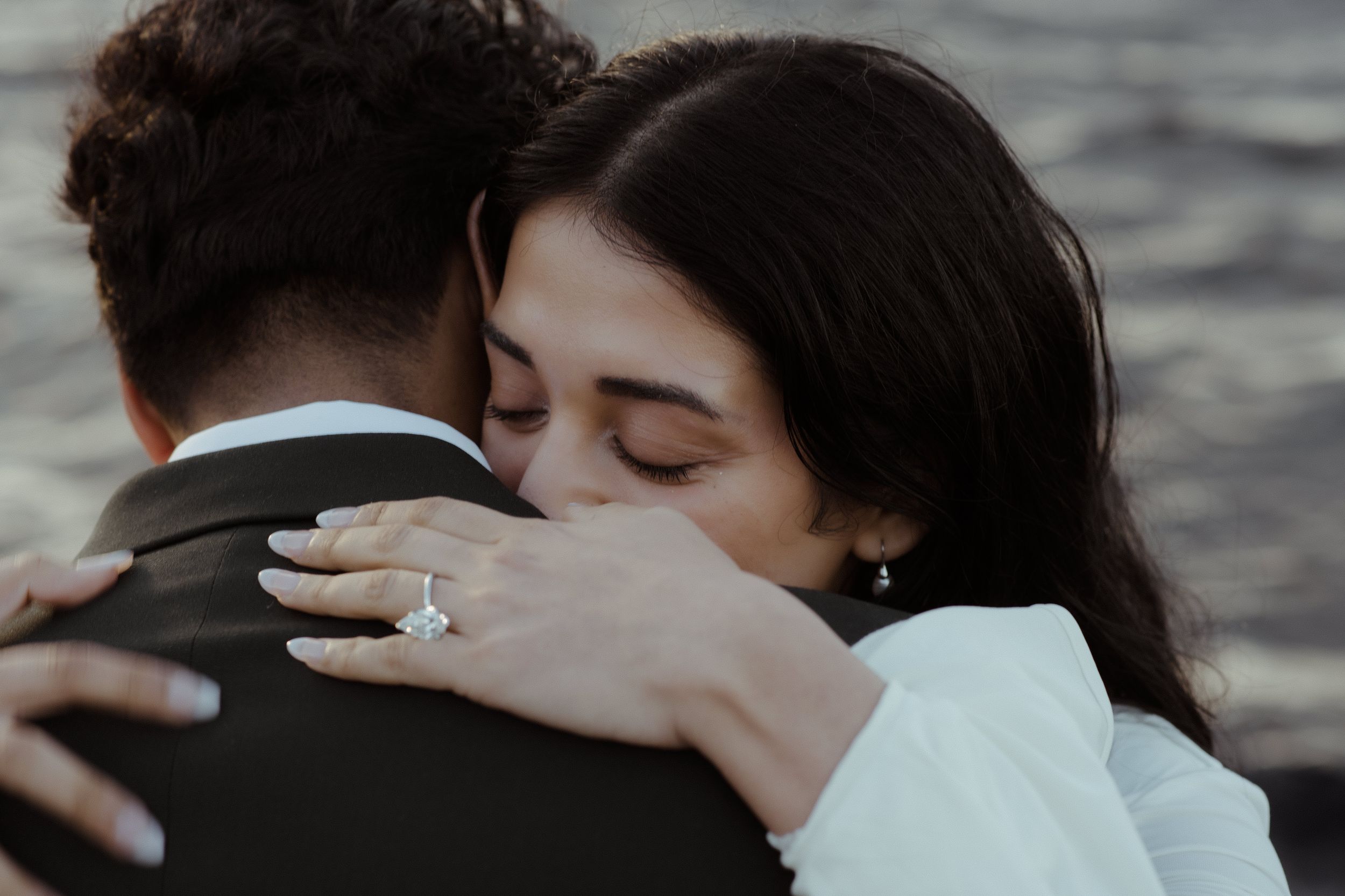 Close up shot of a tender embrace between newlyweds with engagement ring visible near water.