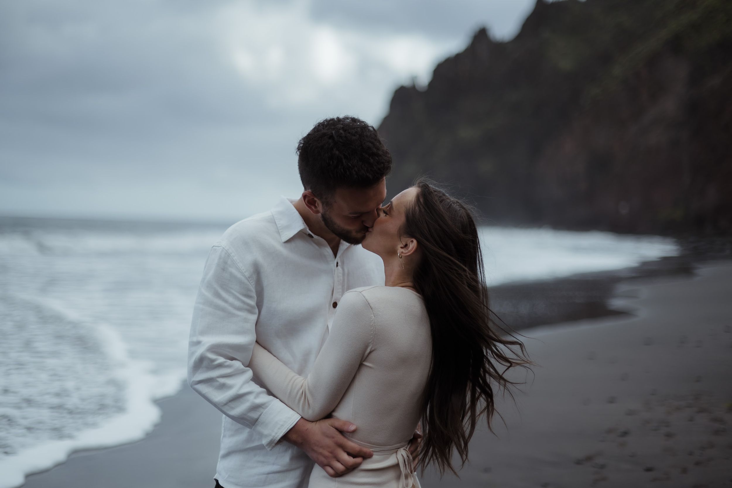 Couple sharing a romantic kiss on a black sand beach with dramatic cliffs and moody ocean waves in the background.