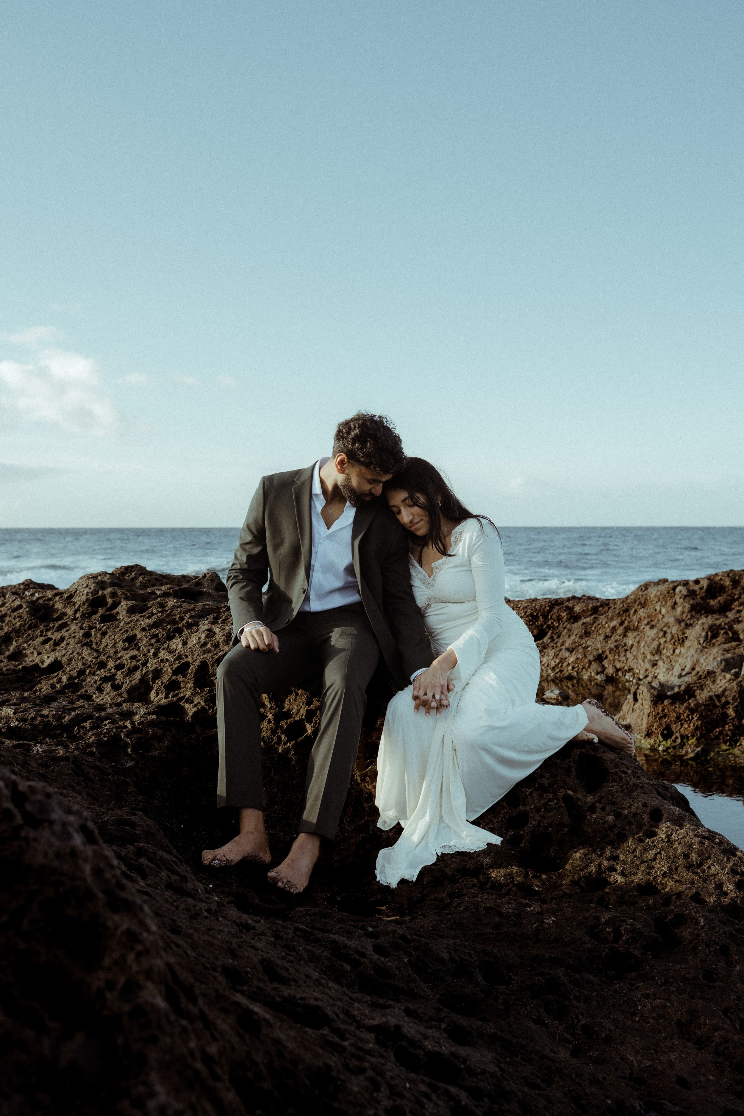 A couple in wedding attire sits together on coastal rocks overlooking the ocean at sunset.