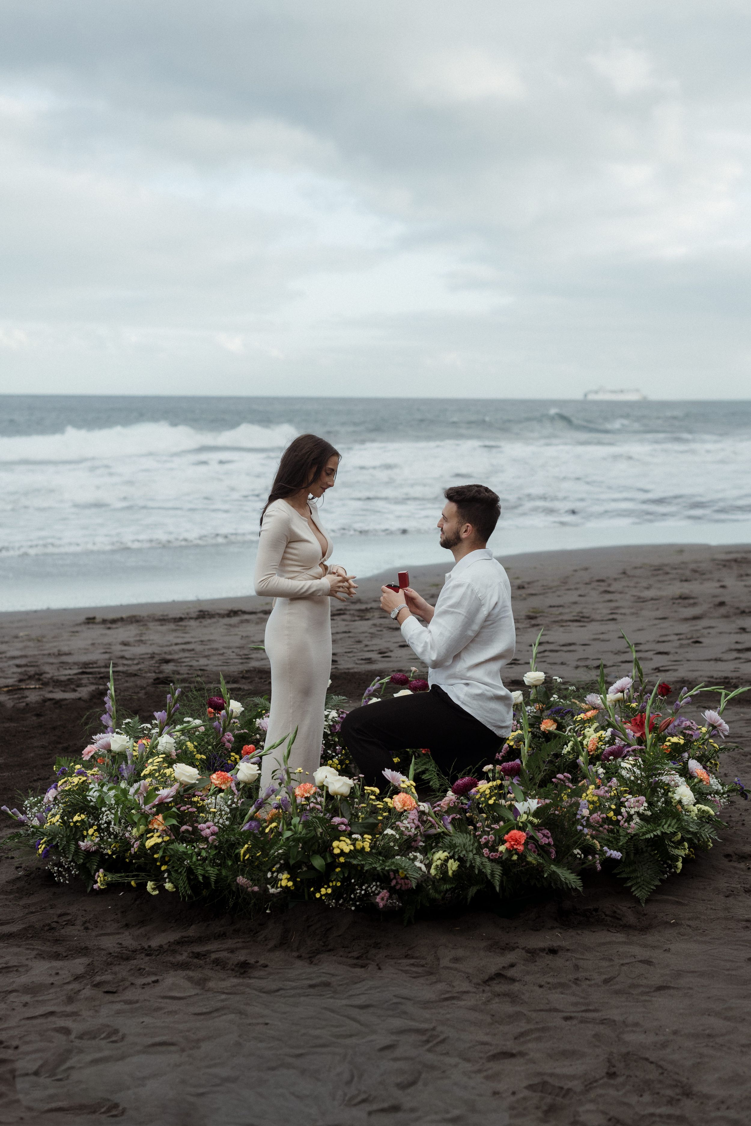 Man proposes to woman on black sand beach surrounded by floral arrangement with ocean backdrop.