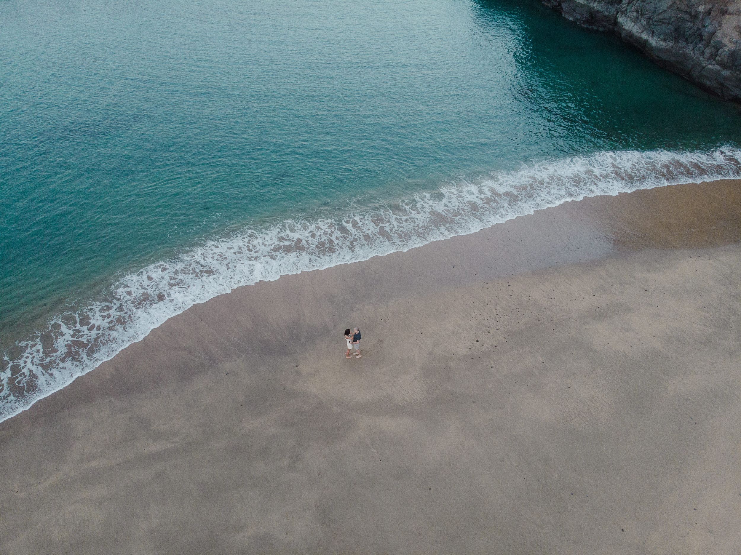 Aerial view of a couple walking on a sandy beach with turquoise ocean waves gently lapping the shore.