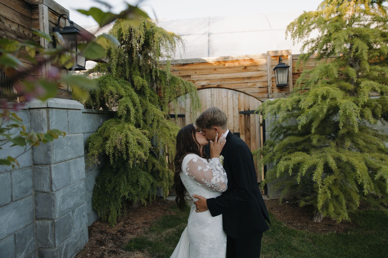 Bride and groom sharing a kiss in a garden setting with lush greenery and wooden gate backdrop.