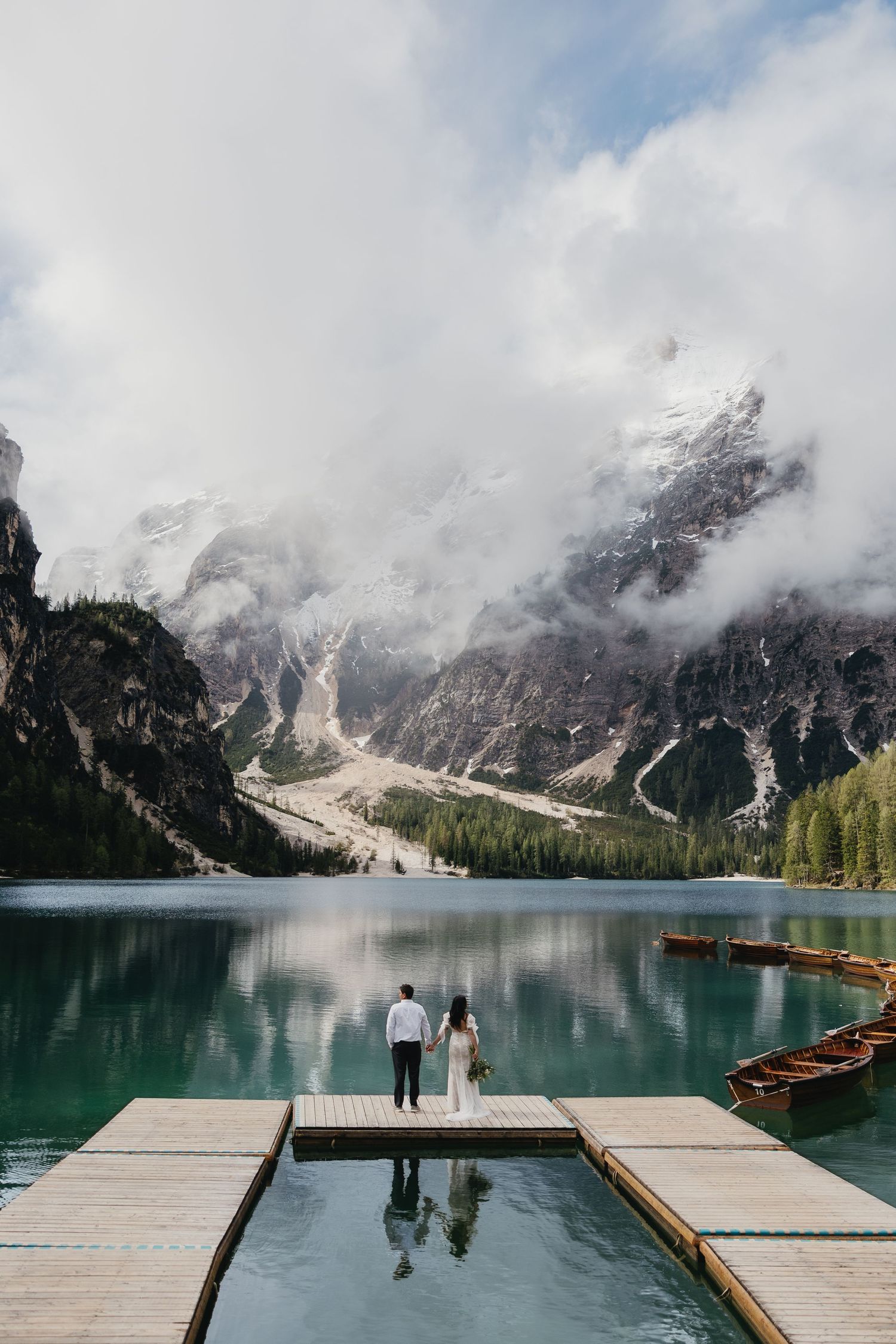 Scenic panorama of Lago di Braies in the Dolomites with wooden pier and boats against misty mountain backdrop.