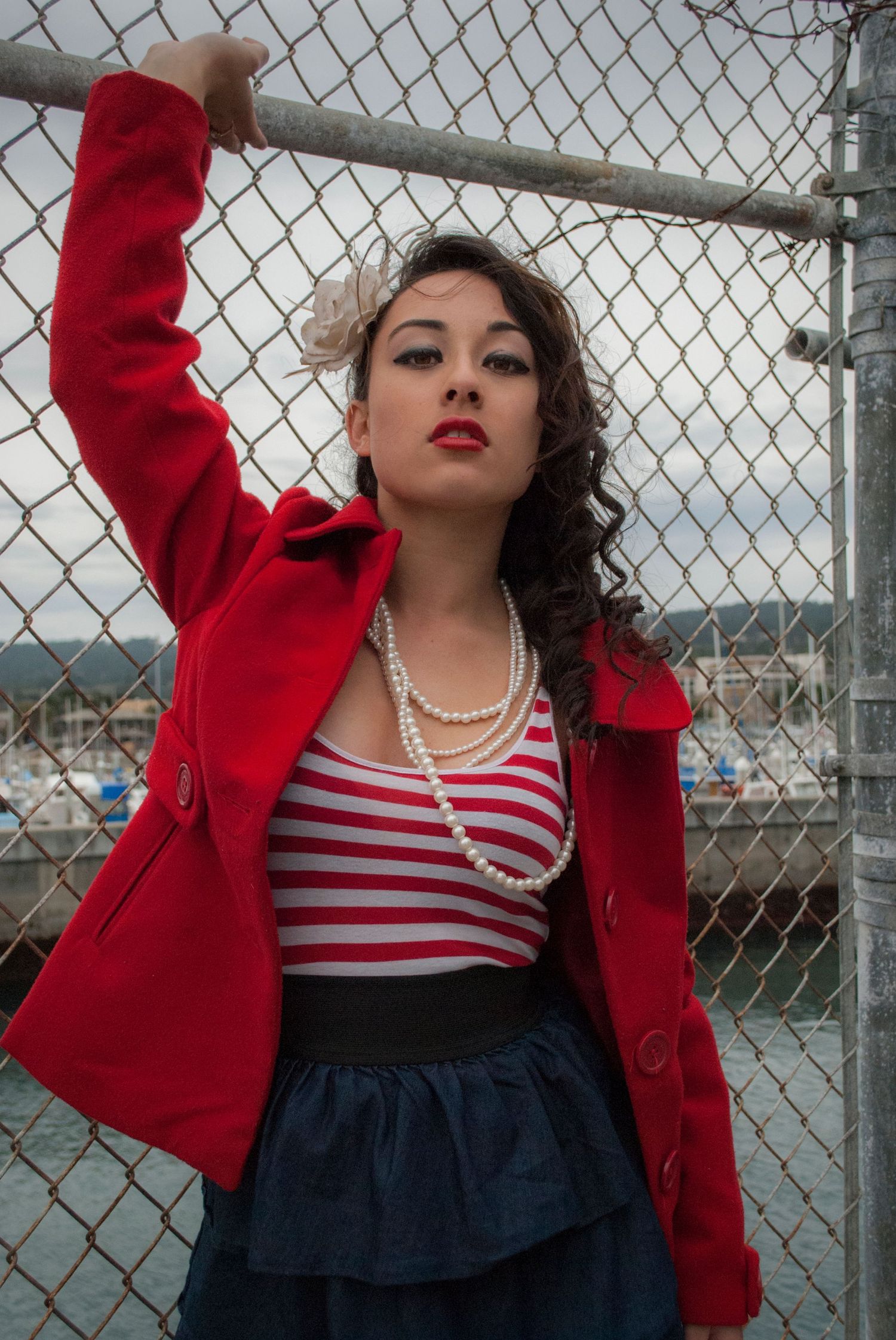 Person in vintage-inspired outfit with red blazer, striped top and navy skirt poses against chain link fence backdrop.