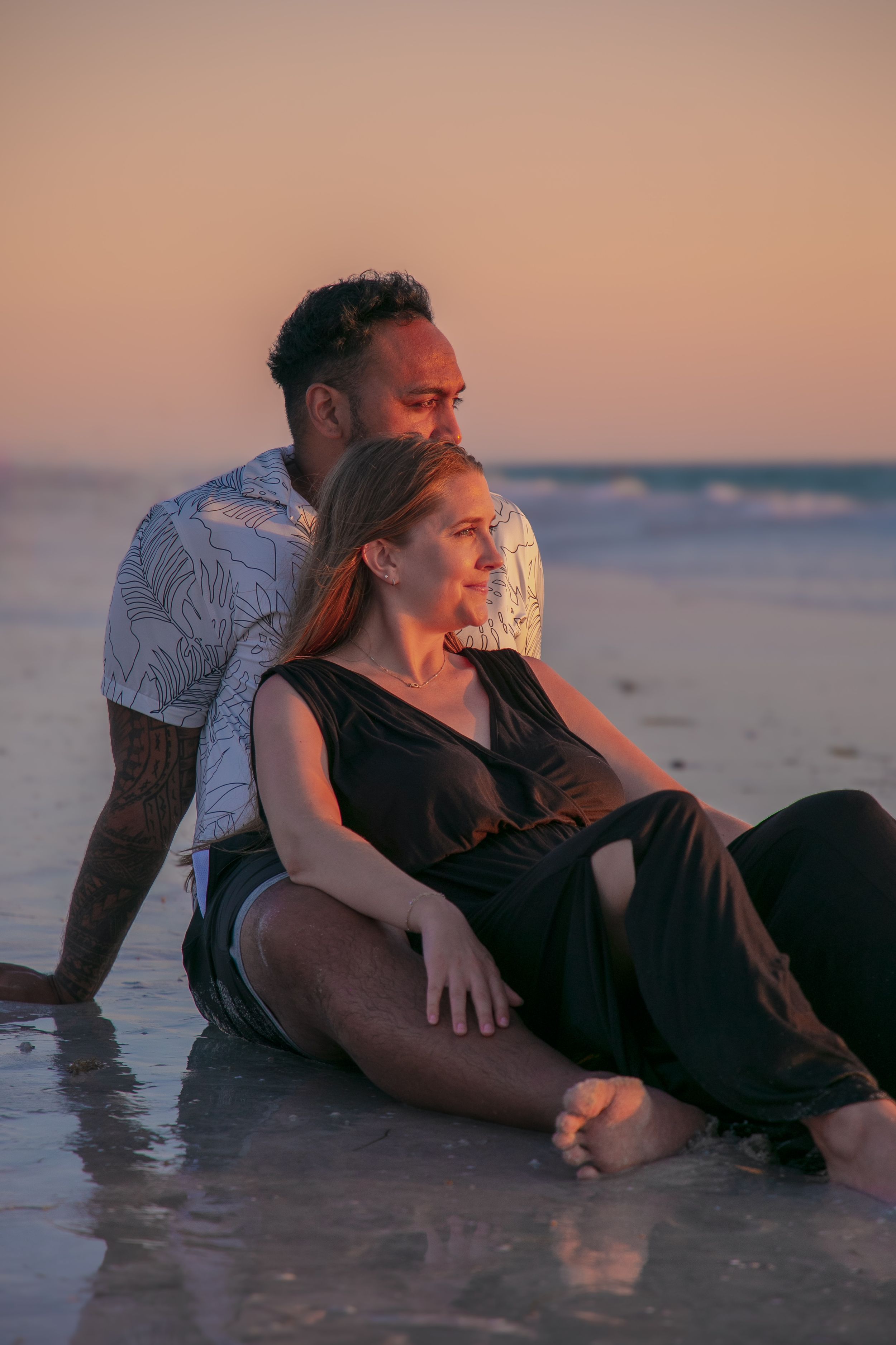 A couple sits together on the beach during a pastel sunset.