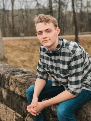A casual portrait of a young person in plaid shirt sitting on stone wall outdoors.