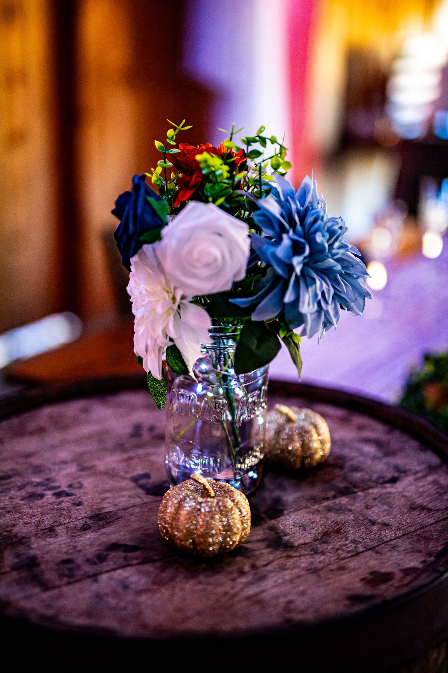 Mason jar centerpiece with blue and white flowers surrounded by small decorative pumpkins.