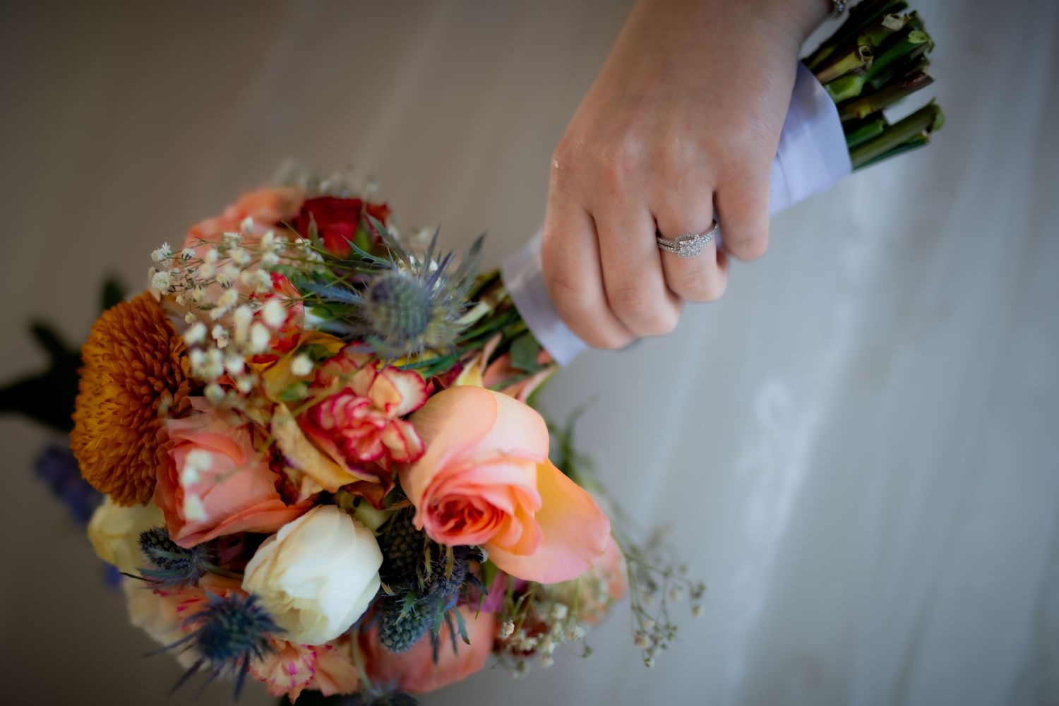 A bridal bouquet featuring coral roses, white flowers and blue thistles with a ring on display.