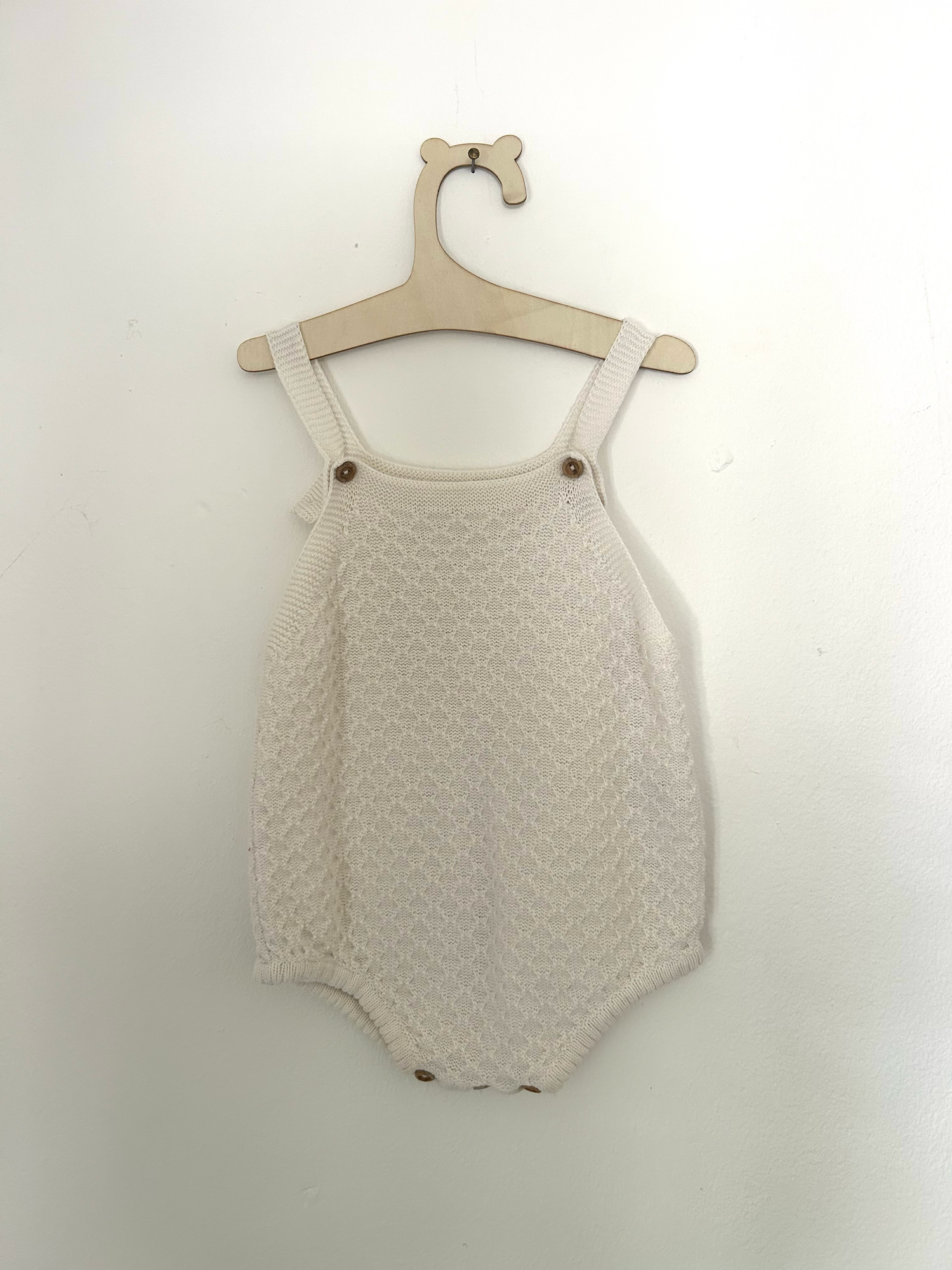 Vintage-style beige knitted baby romper with button details and side pockets hanging on display.