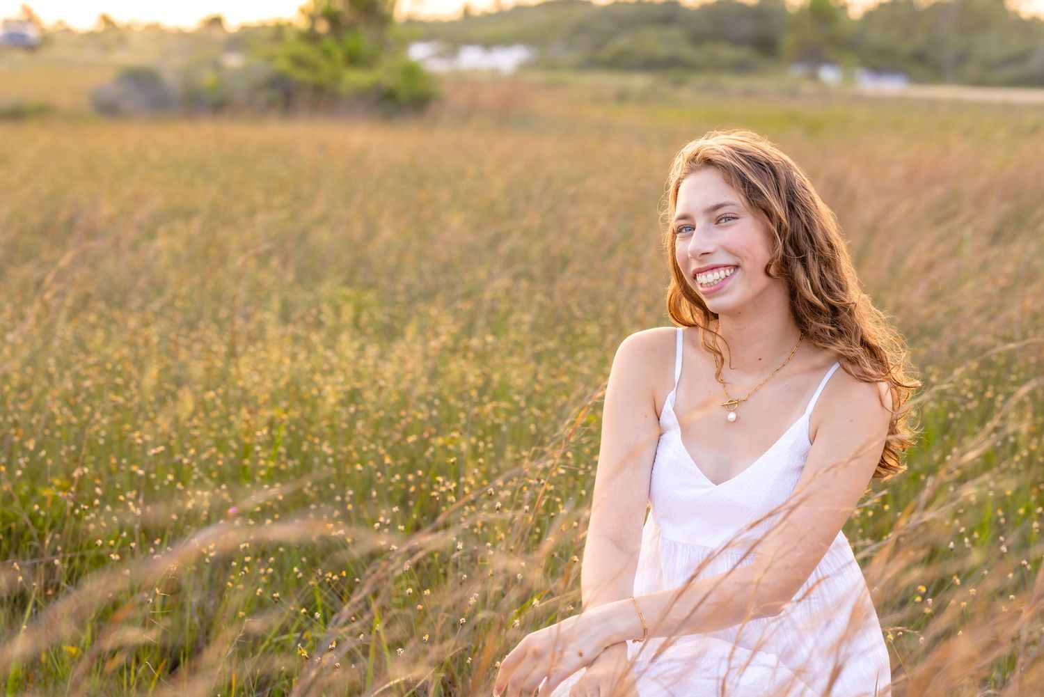 Woman in white dress sits in golden grassy field at sunset, laughing joyfully amid wildflowers.