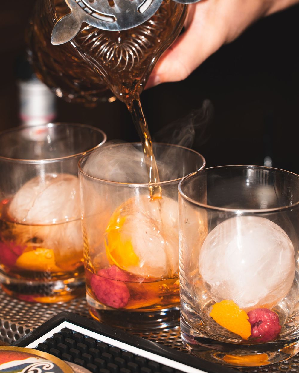 Cocktail being poured over ice in multiple glasses on a dark bar counter.