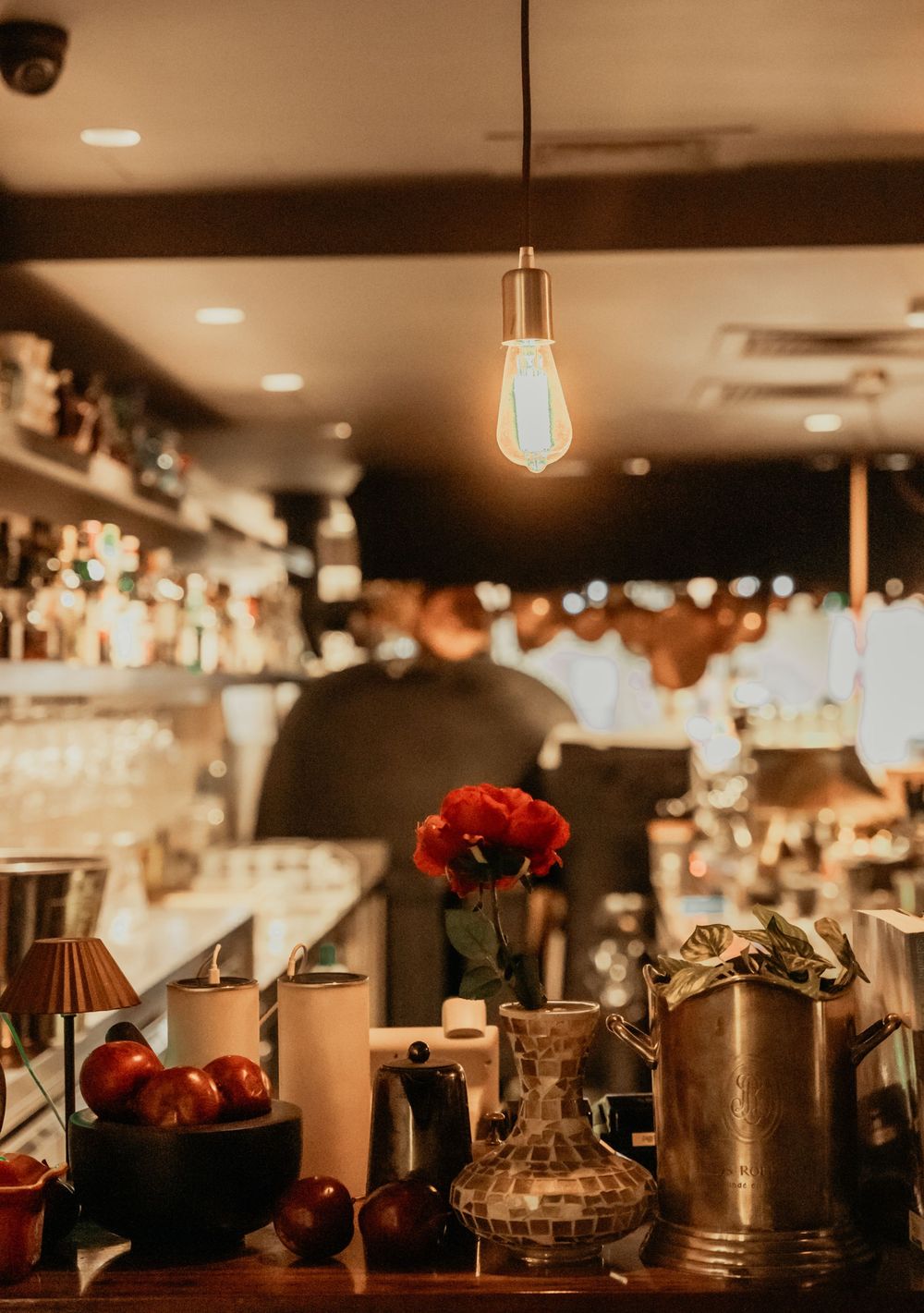 Warm lit restaurant counter with vintage lighting, red roses in vase, and various kitchen items creating cozy ambiance.