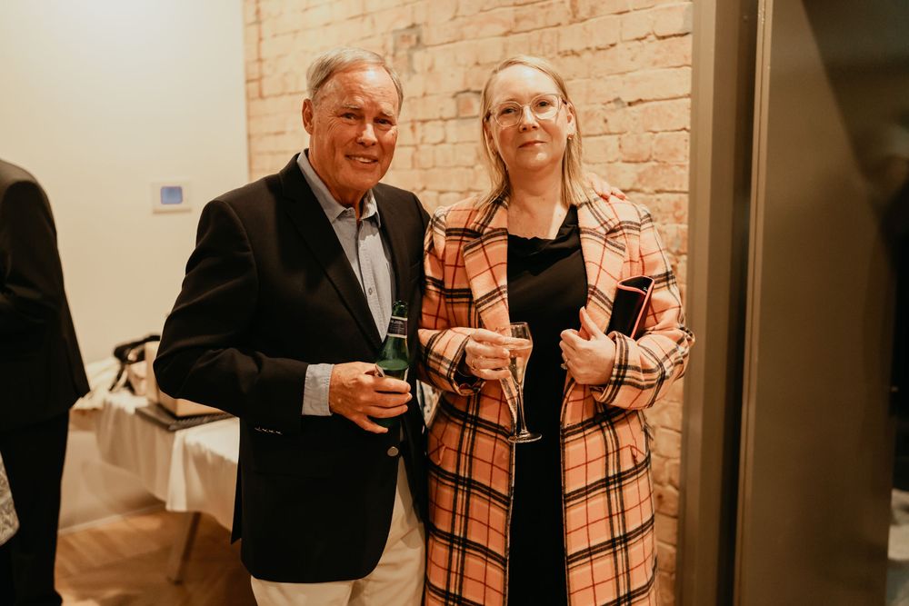 Two people in formal attire stand together smiling and holding wine glasses at an indoor reception.
