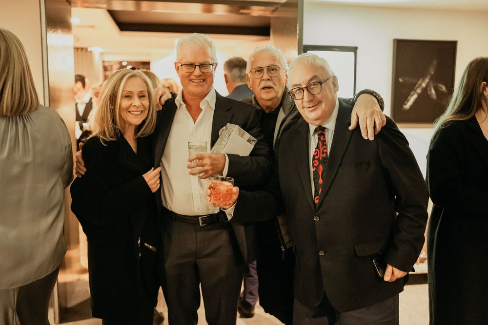 A group of people gather together at an indoor social event with warm lighting and framed artwork on the wall.