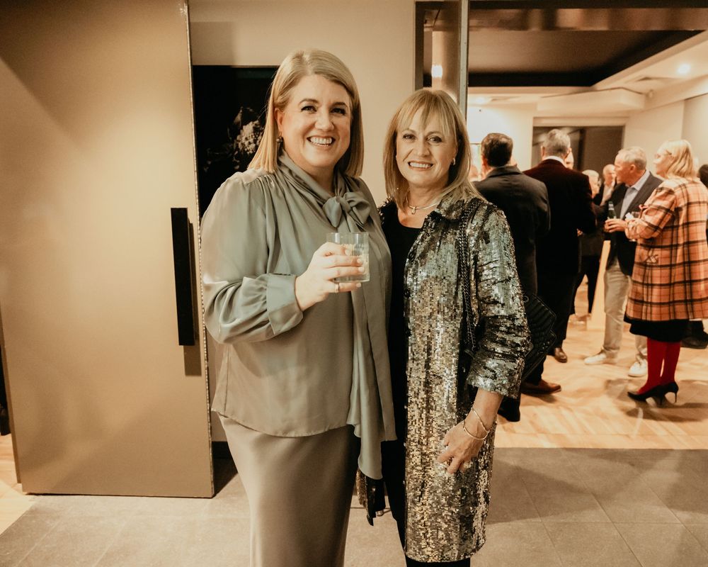 Two women smile while socializing at an indoor gathering, one wearing a beige top and one in a patterned jacket.