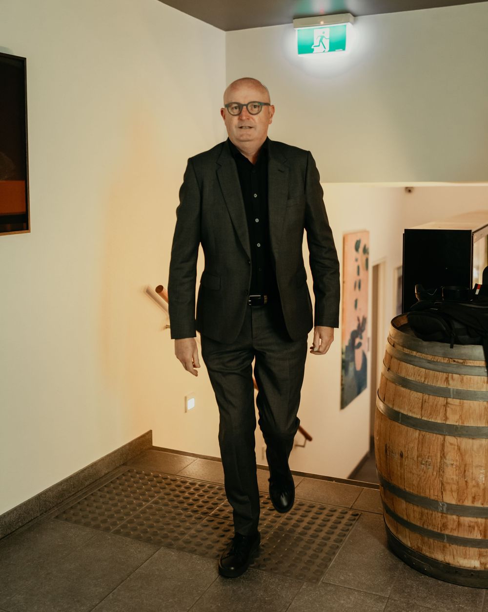 Professional in all-black attire walking past a wooden barrel in a modern interior hallway with exit signage.