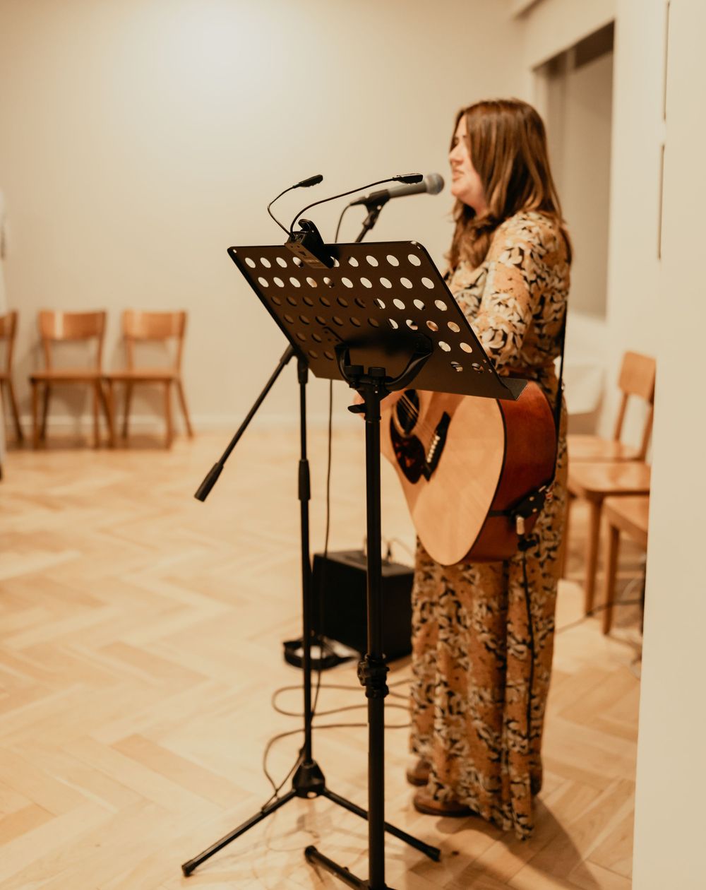 A singer performs in a brown floral dress at a music stand in a well-lit room with wooden flooring.