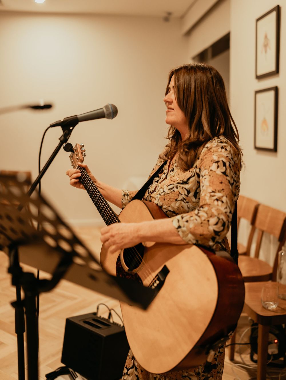 A musician in a floral dress performs with an acoustic guitar and microphone in a warmly lit venue.
