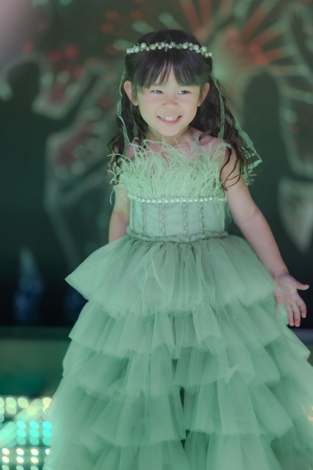 Child wearing a mint green tiered tulle dress and floral crown posing against a sparkly background.