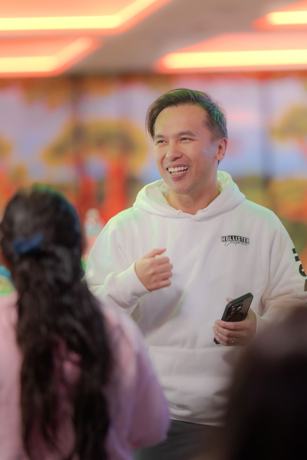 Person wearing white sweatshirt reacts with amusement while looking at phone in warmly lit indoor setting.