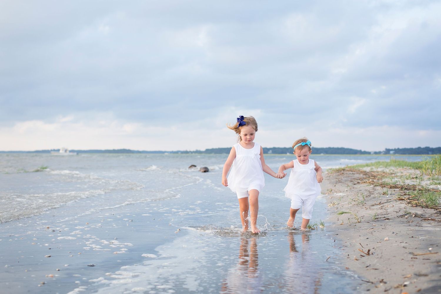 Family wearing white walks along a scenic beach shoreline with gentle waves and cloudy skies in the background.